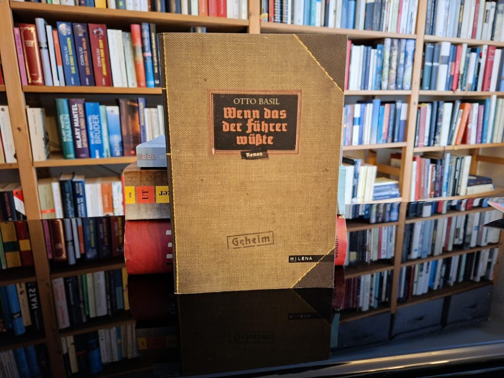Vintage-Buchcover von 'Wenn das der Führer wüsste' von Otto Basil, das in einem Bücherregal steht. Das Buch hat ein beigefarbenes Leinen-Einbanddesign mit einem schwarzen Etikett, auf dem der Titel in gotischer Schrift gedruckt ist. Im Hintergrund sind gefüllte Bücherregale mit einer Vielzahl von Büchern zu sehen.