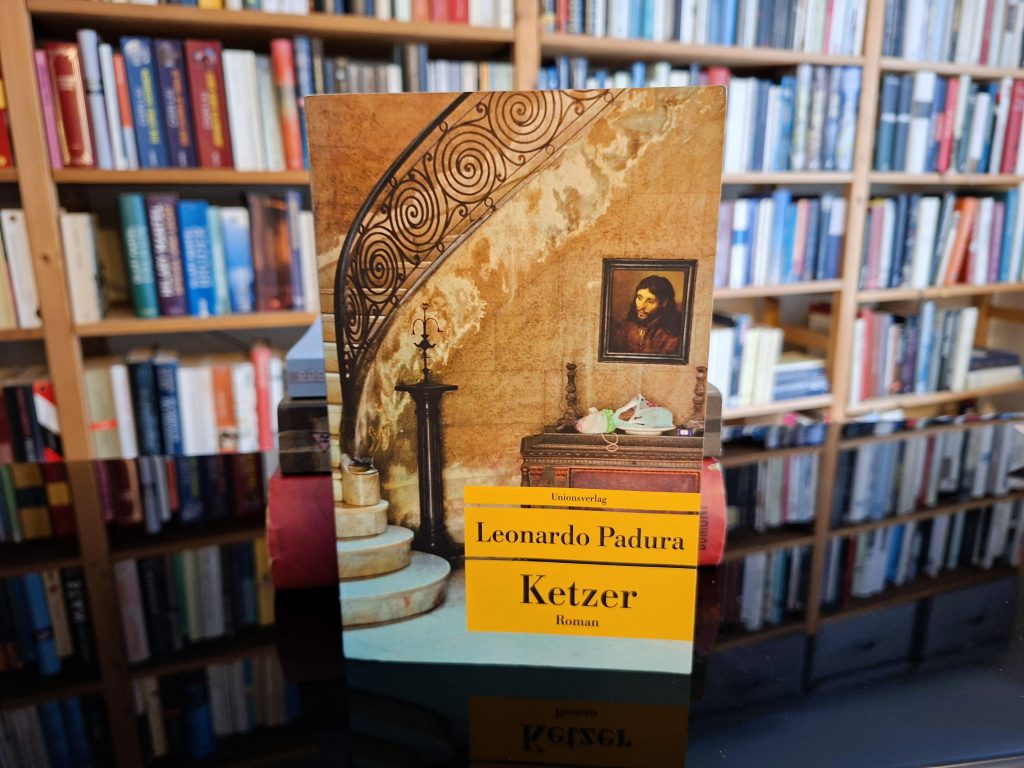 Buchcover von 'Ketzer', einem Roman von Leonardo Padura, das in einem Bücherregal steht. Das Cover zeigt eine künstlerische Darstellung einer alten Treppe mit schmiedeeisernem Geländer und einem kleinen Tisch, auf dem ein gerahmtes Porträt und ein Paar Turnschuhe liegen. Der Titel 'Ketzer' ist auf einem gelben Hintergrund gedruckt.