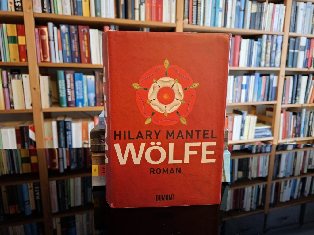 "Buchcover von 'Wölfe', einem Roman von Hilary Mantel, das in einem Bücherregal steht. Das Cover ist in einem kräftigen Rotton gehalten und zeigt eine stilisierte, weiße Tudor-Rose. Der Titel 'Wölfe' ist in großen, weißen Buchstaben gedruckt, darunter steht der Name der Autorin und das Wort 'Roman'. Im Hintergrund sind gefüllte Bücherregale mit einer Vielzahl von Büchern zu sehen.